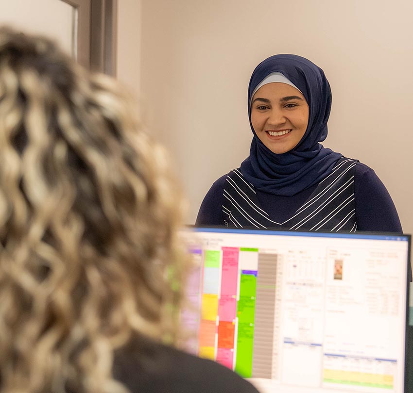 A woman in a hijab smiles while standing near another person who is sitting at a desk with a colorful computer screen.