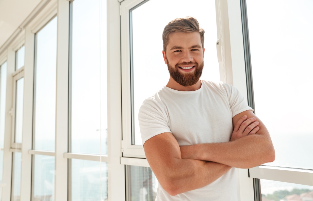 Happy bearded man with crossed arms standing near the window at home and looking at the camera