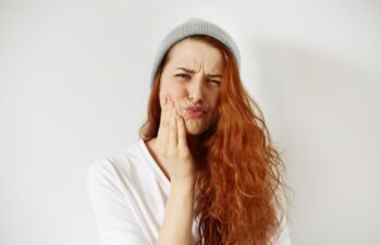 Headshot of redhead teenage woman pressing her cheek with painful expression as if she is having terrible toothache. Negative human emotions face expressions body language. Selective focus