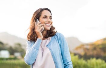 Mature happy woman talking on phone outdoor during sunset. Cheerful hispnic woman using smartphone and looking away. Happy woman in conversation using mobile phone and smiling.