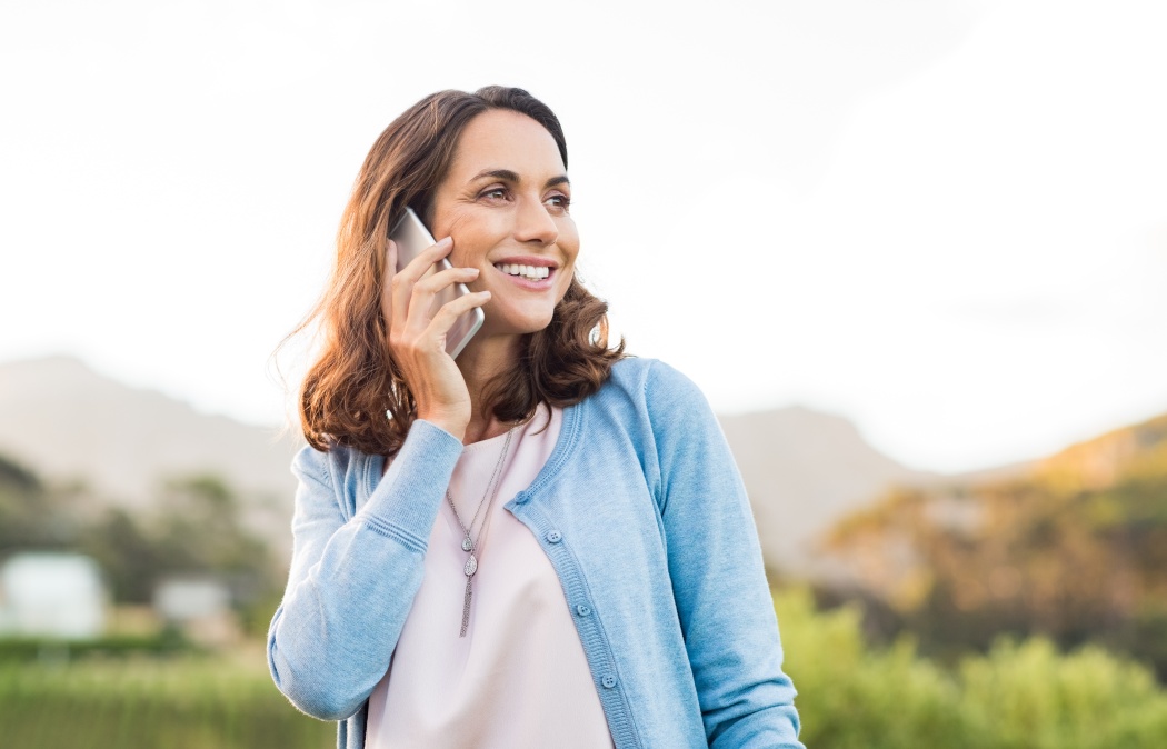 Mature happy woman talking on phone outdoor during sunset. Cheerful hispnic woman using smartphone and looking away. Happy woman in conversation using mobile phone and smiling.