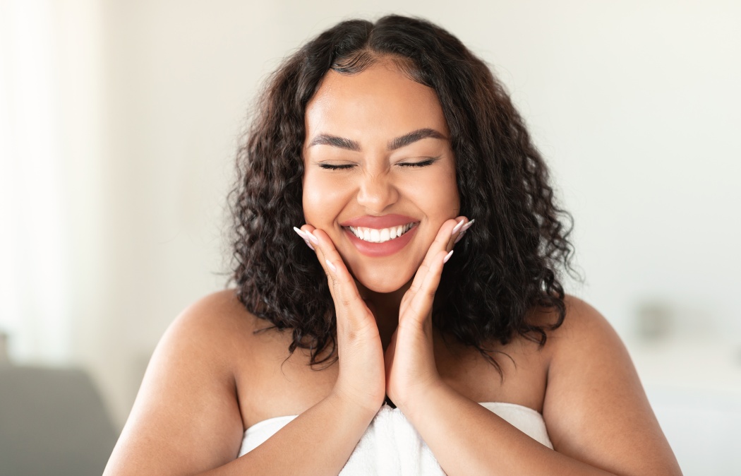 Skin care routine concept. Happy young black plus size woman wrapped in bath towel touching cheeks and smiling