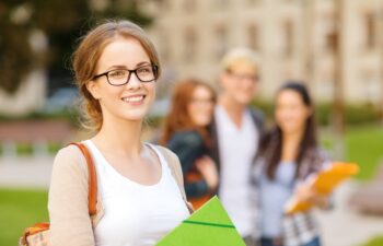 summer holidays, education, campus and teenage concept - smiling female student in black eyeglasses with folders and group in the back