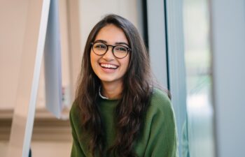 A close up head shot portrait of a preppy, young, beautiful, confident and attractive Indian Asian woman in a green sweater and spectacles in a classroom or office. She is smiling happily.