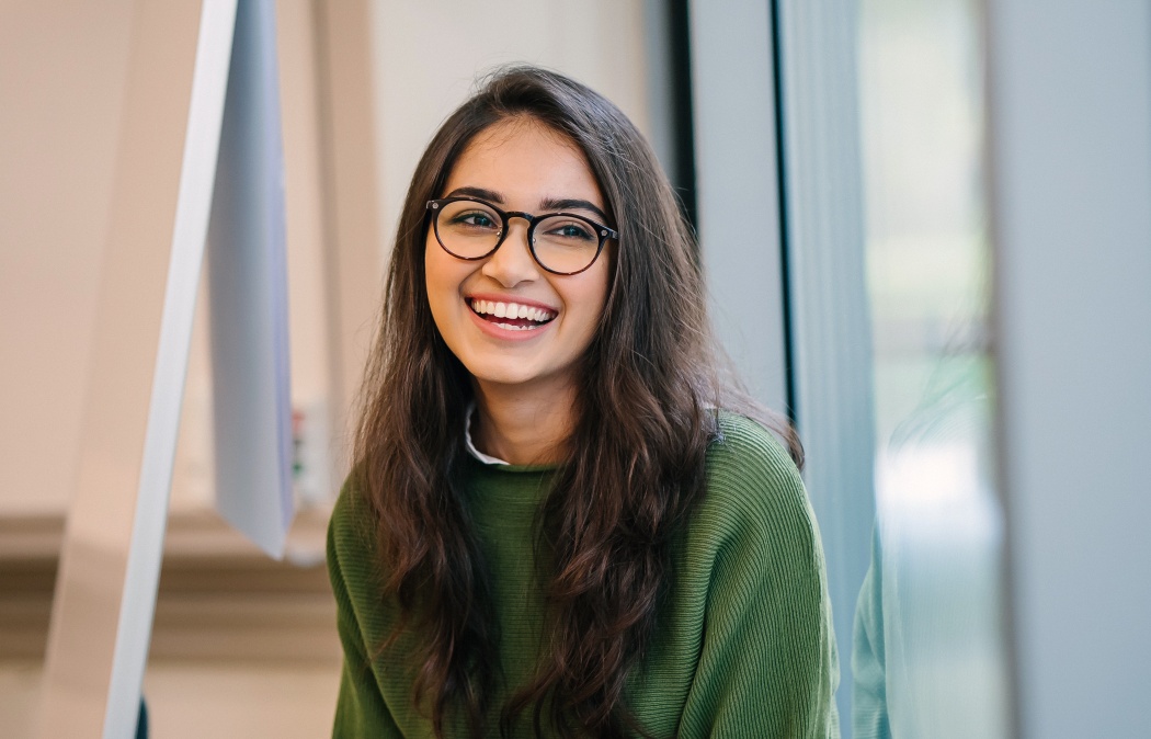 A close up head shot portrait of a preppy, young, beautiful, confident and attractive Indian Asian woman in a green sweater and spectacles in a classroom or office. She is smiling happily.