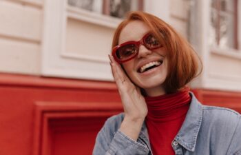 Close-up portrait of young beautiful lady posing outdoors. Pretty red-haired girl wearing turtleneck and blue shirt, adjusting trendy sunglasses and smiling against bright wall background