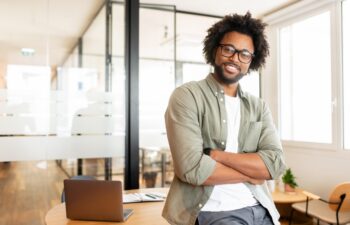 Ambitious highly-skilled handsome curly african-american male employee in glasses and casual wear leaned on office desk and looking at the camera with smile, businessman stands with arms crossed