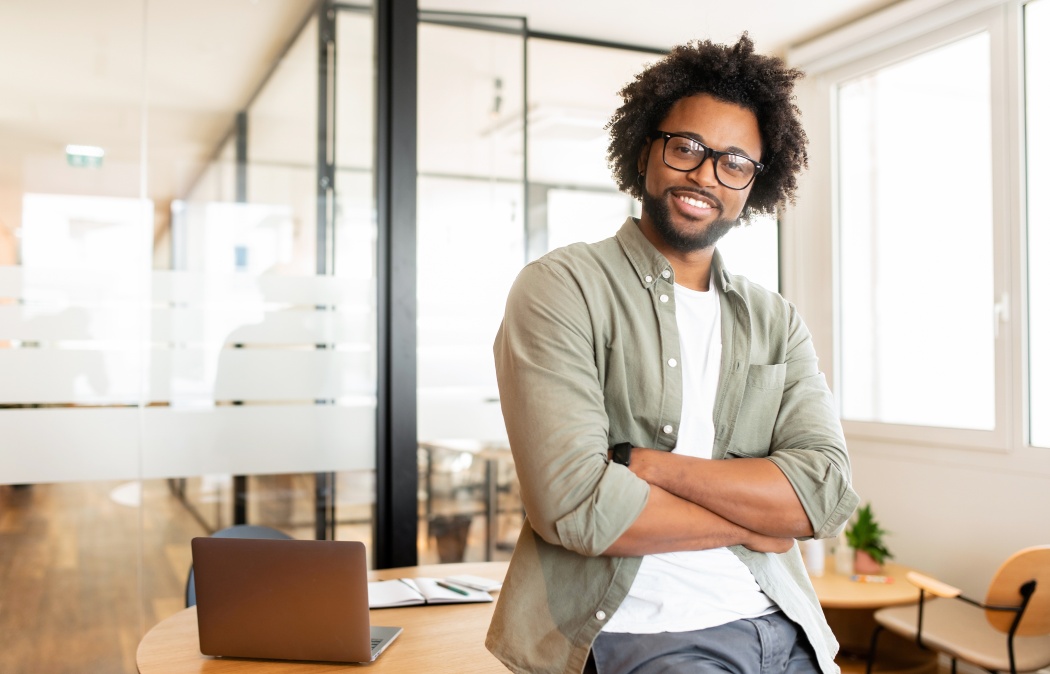 Ambitious highly-skilled handsome curly african-american male employee in glasses and casual wear leaned on office desk and looking at the camera with smile, businessman stands with arms crossed