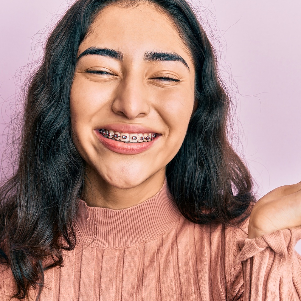 Hispanic teenager girl with dental braces showing orthodontic brackets celebrating achievement with happy smile and winner expression with raised hand