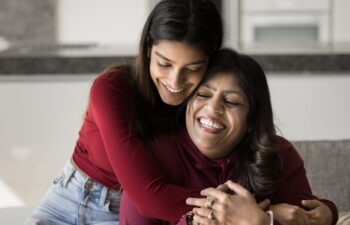 Happy beautiful young Indian daughter woman embracing senior mom from behind with love, tenderness. Cheerful mother and adult kid hugging with closed eyes on home couch, smiling, laughing