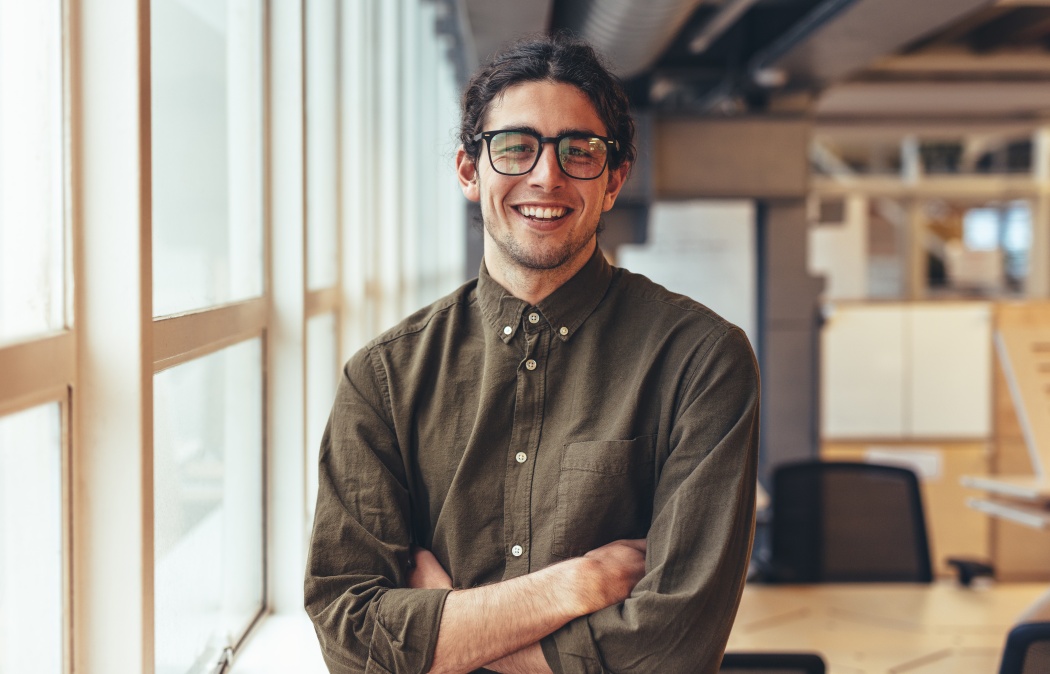 Businessman standing beside a window in office with arms crossed. Portrait of a smiling man standing in office beside a window.